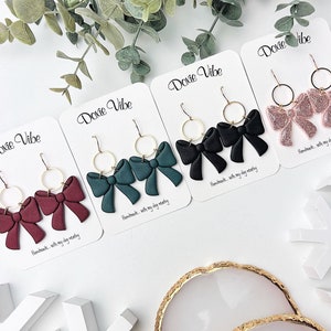 May include: Four pairs of handmade bow earrings on white cards with the text "Doxie Vibe" and "Handmade with my day nearby". The earrings are in four colors: burgundy, green, black, and pink.