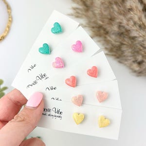 May include: A set of six pairs of heart-shaped earrings. The earrings are made of resin and have a glitter finish. The colors of the earrings are pink, coral, light pink, teal, and gold.
