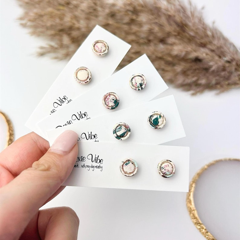 May include: A collection of stud earrings with floral designs, displayed on white cards. The earrings feature gold-colored metal rims and various patterns with pink, green, and white colors. The cards have the text "Xoxie Vibe" printed on them.