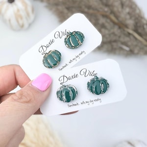 Pu&ograve; includere: Due paia di orecchini a bottone a forma di zucca verde con finiture color oro, presentati su biglietti bianchi. I biglietti riportano il testo "Doxie Vibe" e "Handmade... with my dog nearby".
