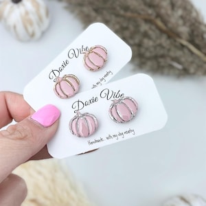 Pu&ograve; includere: Orecchini a bottone a forma di zucca rosa con dettagli in metallo color argento. Gli orecchini sono esposti su biglietti bianchi con il testo "Doxie Vibe" e "Handmade... with my dog nearby."