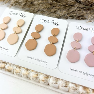 May include: Three pairs of dangle earrings with three different colors: light pink, light brown, and beige. Each earring has three irregular shaped pieces. The earrings are displayed on white cards with the text "Doxie Vibe" and "Handmade... with my dog nearby".