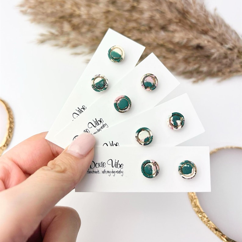 May include: A hand holds several white cards displaying small, round stud earrings. The earrings feature a marbled design with shades of green, pink, and gold. The cards have the text "Doxie Vibe" printed on them.
