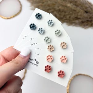 Könnte beinhalten: Eine Auswahl an Ohrsteckern in Pfotenform in verschiedenen Farben, darunter Blau, Grau, Beige und Rosa. Die Ohrringe sind auf weißen Karten präsentiert, mit dem Text "oxie Vibe". Die Ohrringe sind klein und leicht.