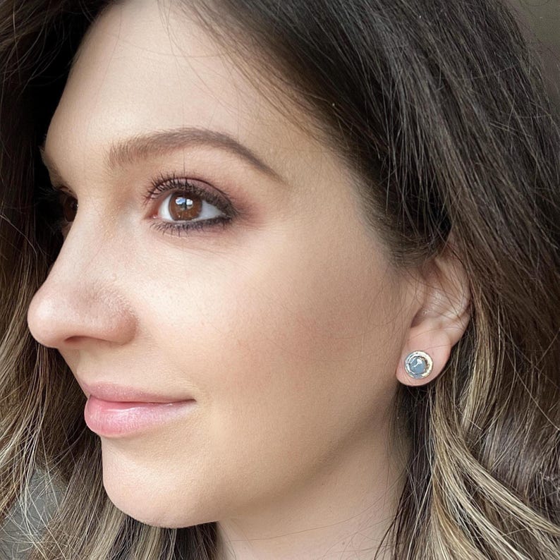 Peut inclure: Gros plan sur le visage d'une personne aux cheveux et aux yeux bruns. La personne porte de petites boucles d'oreilles rondes avec une pierre bleu clair et un sertissage argent&eacute;.