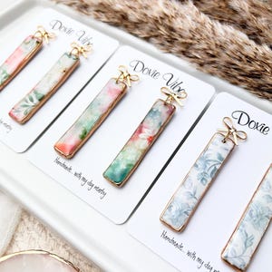 May include: Three pairs of rectangular drop earrings with gold-toned edges, each featuring a unique floral pattern in soft pastel colors. Each earring is attached to a white display card labeled Doxie Vibe with a small gold-toned bow accent.