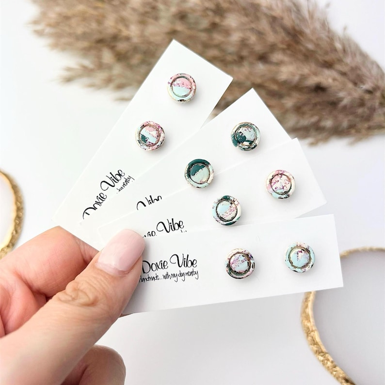 May include: A collection of small, round stud earrings with a marbled design in shades of pink, green, and blue. The earrings are displayed on white cards with the text "Doxie Vibe" printed on them. The earrings are held by a hand.