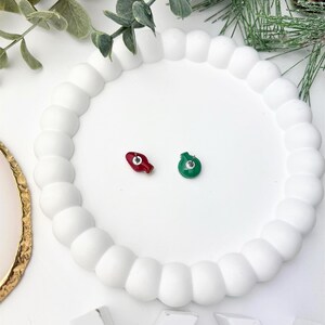May include: Two Christmas light bulb stud earrings, one red and one green, rest on a white, round, beaded tray. The earrings are small and detailed, perfect for holiday wear. The tray is surrounded by greenery.