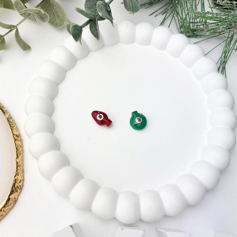 May include: Two Christmas light bulb stud earrings, one red and one green, rest on a white, round, beaded tray. The earrings are small and detailed, perfect for holiday wear. The tray is surrounded by greenery.