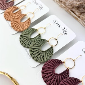May include: Three pairs of handmade polymer clay earrings. The earrings are in the shape of a fan with a gold hoop. The colours are terracotta, olive green, and burgundy.