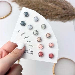 Peut inclure: Collection de boucles d'oreilles &agrave; clous de diff&eacute;rentes couleurs, dont gris, bleu clair et rose, pr&eacute;sent&eacute;es sur des cartes blanches. Chaque boucle d'oreille a une bordure argent&eacute;e. Le texte "Doxie Vibe" est visible.