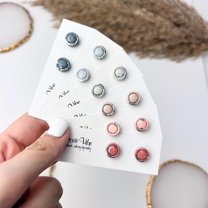 Peut inclure: Collection de boucles d'oreilles &agrave; clous de diff&eacute;rentes couleurs, dont gris, bleu clair et rose, pr&eacute;sent&eacute;es sur des cartes blanches. Chaque boucle d'oreille a une bordure argent&eacute;e. Le texte "Doxie Vibe" est visible.