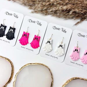 May include: A collection of handmade cat-shaped earrings in black, hot pink, white, and light pink. Each pair is displayed on a white card with the text "Doxie Vibe" and "Handmade... with my dog nearby."