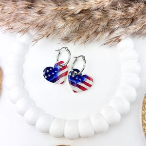 May include: A pair of silver hoop earrings featuring heart-shaped charms with a red, white, and blue American flag pattern. The charms have a glossy finish and are suspended from thin, circular silver-toned metal hoops with a secure clasp.