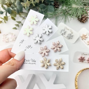 May include: A collection of snowflake stud earrings in various colors, including iridescent, silver, rose gold, and gold. Each pair is displayed on a white card with the text "Doxie Vibe" and "handmade with my dog besty".