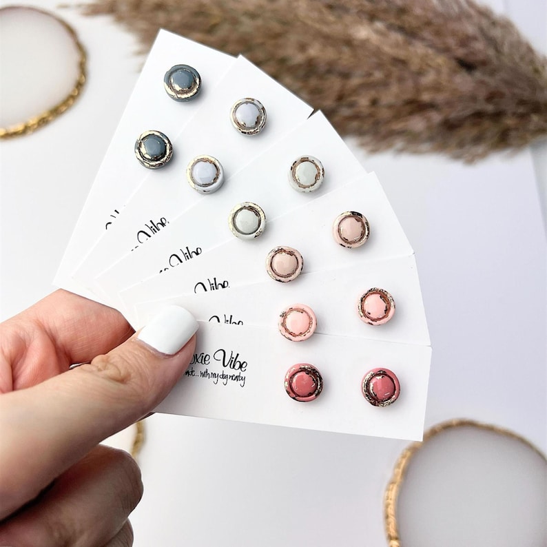 Peut inclure: Collection de boucles d'oreilles &agrave; clous de diff&eacute;rentes couleurs, dont gris, bleu clair et rose, pr&eacute;sent&eacute;es sur des cartes blanches. Chaque boucle d'oreille a un bord m&eacute;tallique. Les cartes sont tenues par une main avec un ongle peint en blanc. Le texte "Pixie Vibe" est visible.
