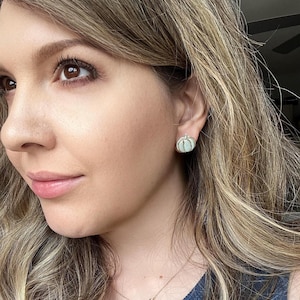 Pu&ograve; includere: Primo piano del viso di una persona con un orecchino a bottone a forma di zucca azzurra. L'orecchino &egrave; realizzato in materiale azzurro con dettagli in metallo color argento. La persona ha lunghi capelli biondi ondulati.