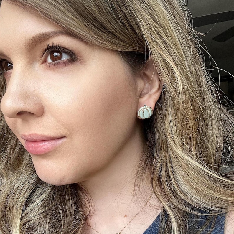 Pu&ograve; includere: Primo piano del viso di una persona con un orecchino a bottone a forma di zucca azzurra. L'orecchino &egrave; realizzato in materiale azzurro con dettagli in metallo color argento. La persona ha lunghi capelli biondi ondulati.