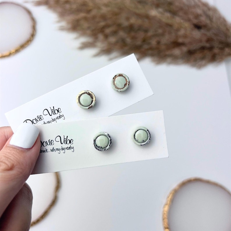 Peut inclure: Deux paires de boucles d'oreilles &agrave; clous sur des cartes blanches. Chaque boucle d'oreille a un centre vert clair avec une bordure m&eacute;tallique. Les cartes indiquent "Doxie Vibe...with my day nasty". Une paire a une bordure dor&eacute;e, l'autre une bordure argent&eacute;e.