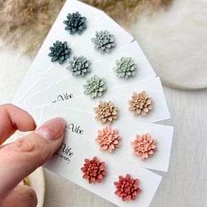 Op de afbeelding: Een verzameling bloemenknopjes in verschillende kleuren, waaronder teal, saliegroen, perzik en rood. Elk paar wordt gepresenteerd op een witte kaart met het woord "Vibe" erop gedrukt. De oorbellen zijn in rijen gerangschikt.