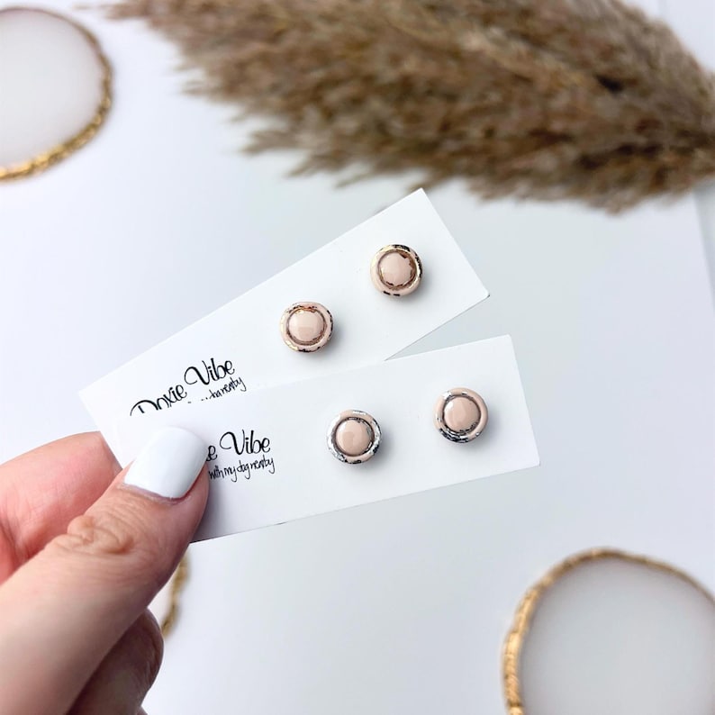 Peut inclure: Boucles d'oreilles &agrave; clous avec un centre rose clair et une bordure argent&eacute;e. Elles sont pr&eacute;sent&eacute;es sur des cartons blancs avec le texte "Dixie Vibe" en &eacute;criture manuscrite. Une main avec du vernis &agrave; ongles blanc tient les boucles d'oreilles.