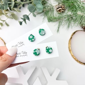 May include: Green Christmas ornament earrings with white snowflake designs. The earrings are on white cards with the text "Doxie Vibe" and a cursive tagline. Festive holiday jewelry.