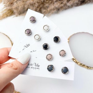 May include: A collection of stud earrings in various colors, including gray, black, pink, and white, with silver-colored metal rims. The earrings are displayed on white cards with the text "Doxie Vibe".