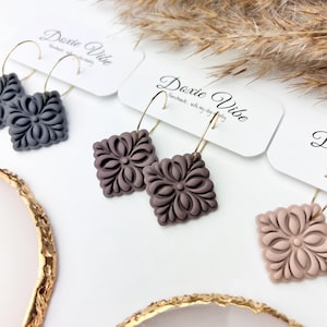 May include: Close-up of several pairs of square floral earrings in various colours, including grey, mauve, and pink. The earrings are attached to gold-coloured hoops. The brand name "Doxie Vibe" is printed on the white earring display cards.