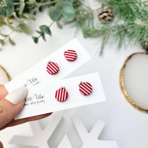 May include: Red and white striped Christmas ornament stud earrings. The earrings are round with a gold-colored top and are displayed on white cards. The cards have the text "oxie Vibe" printed on them.