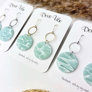 May include: Earrings with a light blue and white marbled design. The earrings have a circular shape and are attached to a gold or silver hoop and hook. The cards read "Dovie Vibe" and "Handmade... with my dog nearby."