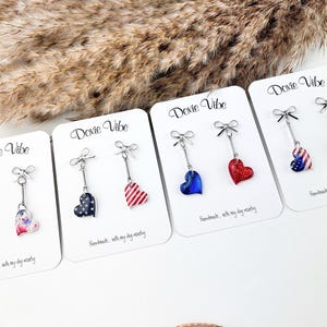 May include: Four pairs of dangle earrings on white display cards labeled Doxie Vibe. Each earring features a silver-tone bow at the top, a thin metal bar, and a heart-shaped charm with various patterns including stars, stripes, and glittery red or blue.
