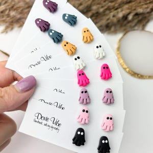 May include: A collection of colorful ghost-shaped earrings in various shades, including purple, teal, orange, white, pink, and black. The earrings are displayed on white cards with the text "Doxie Vibe" and "Handmade...with my dog nearby".