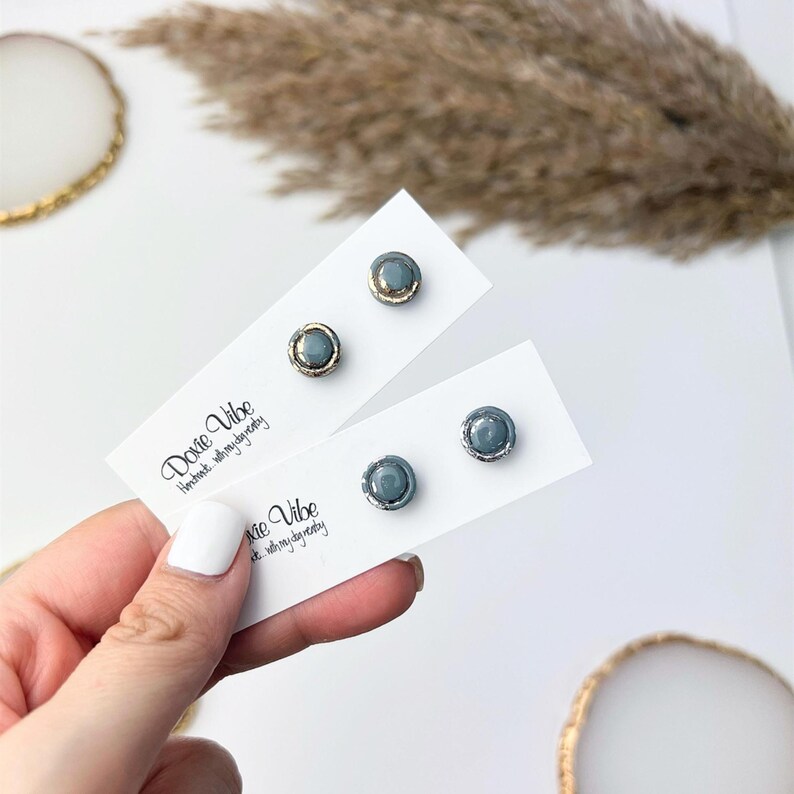 Peut inclure: Deux paires de boucles d'oreilles &agrave; clous avec un centre en pierre grise et une bordure m&eacute;tallique dor&eacute;e ou argent&eacute;e. Les boucles d'oreilles sont pr&eacute;sent&eacute;es sur des cartes blanches avec le texte "Dovie Vibe Handmade with my day today".
