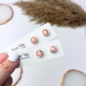 Peut inclure: Quatre boucles d'oreilles &agrave; clous en or rose avec un centre rose clair sont pr&eacute;sent&eacute;es sur des cartes blanches. Les cartes portent l'inscription "Doxie Vibe...jewelry". Les boucles d'oreilles sont rondes avec un petit bord sur&eacute;lev&eacute;.