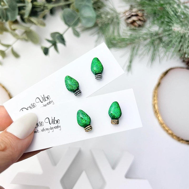 May include: Green glitter Christmas light bulb stud earrings. The earrings are on white cards with the text "Doxie Vibe... heavenly" printed on them. The earrings have silver and gold colored bases.