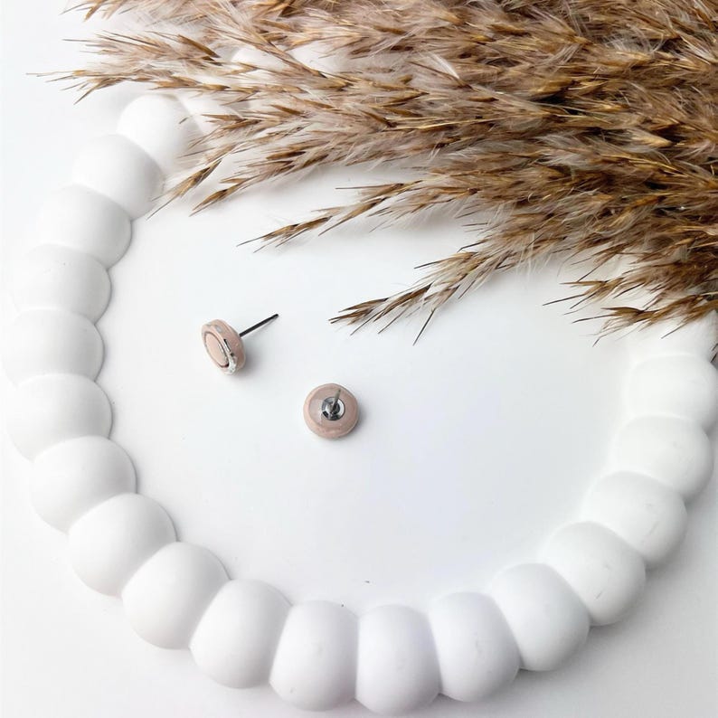 Peut inclure: Deux petites boucles d'oreilles rondes avec un centre rose clair et une bordure argent&eacute;e. Les boucles d'oreilles sont pr&eacute;sent&eacute;es sur une surface blanche textur&eacute;e avec de l'herbe ornementale s&eacute;ch&eacute;e de couleur beige en arri&egrave;re-plan. Bijoux minimalistes.