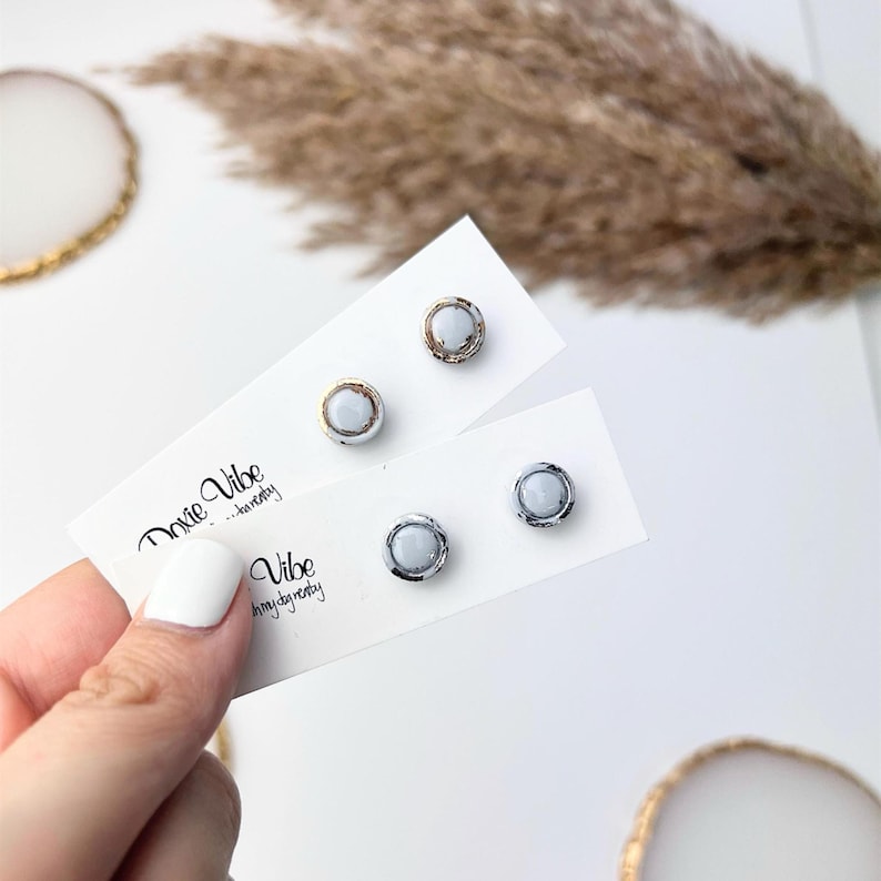 Peut inclure: Boucles d'oreilles &agrave; clous avec une pierre centrale bleu clair et un bord m&eacute;tallique. Une paire a une bordure dor&eacute;e, l'autre une bordure argent&eacute;e. Les boucles d'oreilles sont pr&eacute;sent&eacute;es sur des cartes blanches avec le texte "Doxie Vibe".