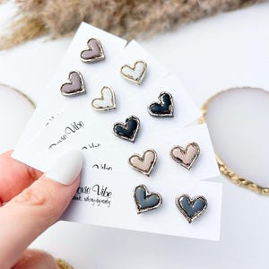 Peut inclure: Boucles d'oreilles clous en forme de cœur de différentes couleurs, dont noir, blanc, gris et beige. Chaque boucle d'oreille a une bordure métallique et est présentée sur une carte blanche. Les cartes portent le texte "oxie Vibe".