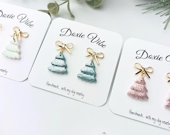 May include: Close-up of handmade Christmas tree earrings in various colors, including green and pink, with gold-colored bow accents. Each earring is displayed on a white card with the text "Doxie Vibe" and "Handmade... with my dog nearby."