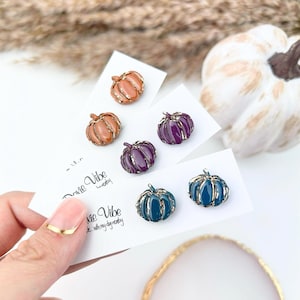 May include: A collection of pumpkin-shaped earrings in various colors, including orange, purple, and teal, with gold-colored trim. The earrings are displayed on white cards with the text "ovie Vibe" and a small pumpkin decoration. A fall-themed accessory.