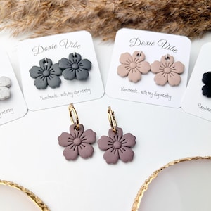 May include: Close-up of handmade flower earrings in various colors, including gray, tan, and mauve. The earrings are displayed on white cards with the text "Doxie Vibe" and "Handmade... with my dog nearby". Gold hoop earrings with mauve flowers are also visible.