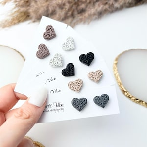 May include: A collection of heart-shaped stud earrings in various colors, including black, white, gray, and beige. Each earring features a geometric floral pattern. The earrings are displayed on white cards with the text "Joxie Vibe".