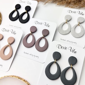 Op de afbeelding: Een collectie handgemaakte druppelvormige oorbellen in diverse kleuren, waaronder zwart, mauve, gespikkeld grijs en oudroze. Elke paar wordt gepresenteerd op een witte kaart met de tekst "Doxie Vibe" en "Handmade... with my dog nearby."