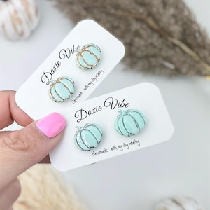 Pu&ograve; includere: Due paia di orecchini a bottone a forma di zucca azzurra con dettagli color argento, esposti su cartoncini bianchi. Le carte presentano il testo "Doxie Vibe" e "Handmade... with my dog nearby."