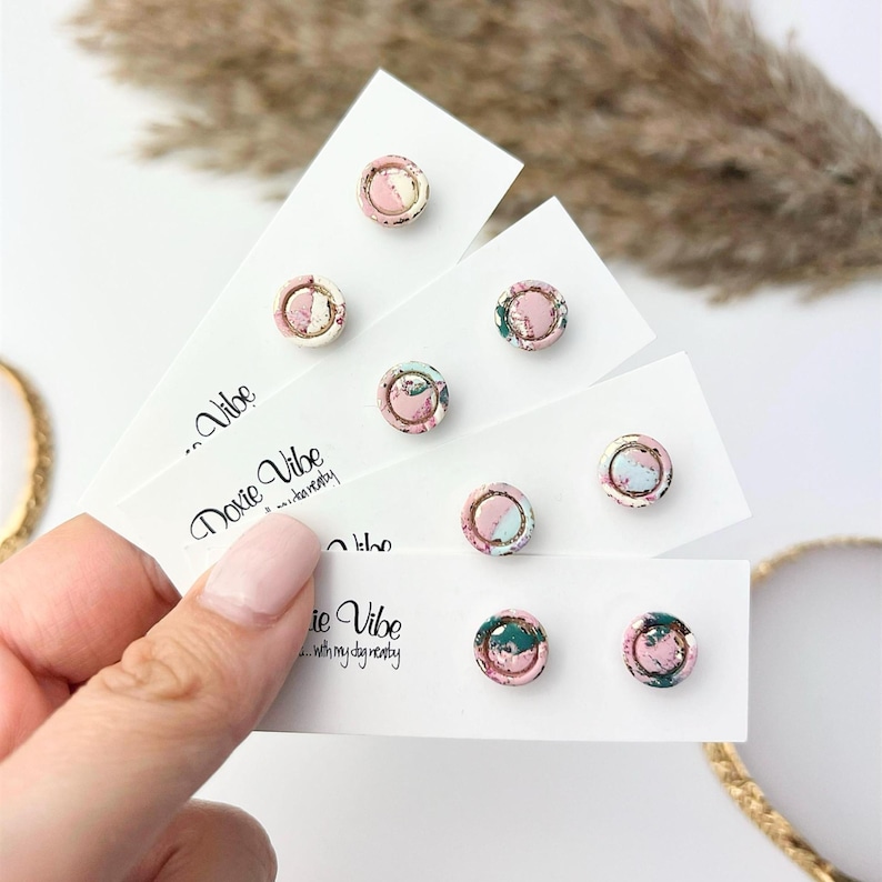 May include: A collection of round stud earrings with a marbled design in pink, green, and blue hues. The earrings are displayed on white cards with the text "Doxie Vibe...with my dog nearby".