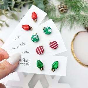 May include: A collection of Christmas-themed stud earrings. The earrings feature designs like red glittery light bulbs, green ornaments with white snowflakes, and red and white striped ornaments. The earrings are displayed on white cards with the text "Pixie Vibe...with my dog nearby."