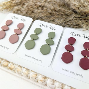 Op de afbeelding: Drie paar hangende oorbellen met drie geometrische vormen op elke oorbel. De oorbellen zijn in drie kleuren: stoffig roze, saliegroen en bordeauxrood.