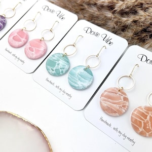 Peut inclure: Plusieurs paires de boucles d'oreilles artisanales présentées sur des cartes blanches. Chaque boucle d'oreille est ornée d'un crochet doré et d'un pendentif circulaire avec un motif marbré dans des tons de rose, de turquoise et de pêche. Le texte "Dovie Vibe" et "Handmade... with my dog nearby" sont visibles.