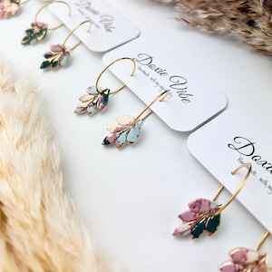 May include: Gold hoop earrings with leaf-shaped pendants in pink, green, and blue. The earrings are displayed on white cards with the text "Doxie Vibe" and are handmade with clay.