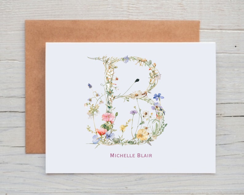 Pu&ograve; includere: Un biglietto bianco con una lettera B floreale realizzata con fiori ad acquerello in tonalit&agrave; di rosa, blu, giallo e verde. Il nome "Michelle Blair" &egrave; stampato in rosa sotto la lettera.